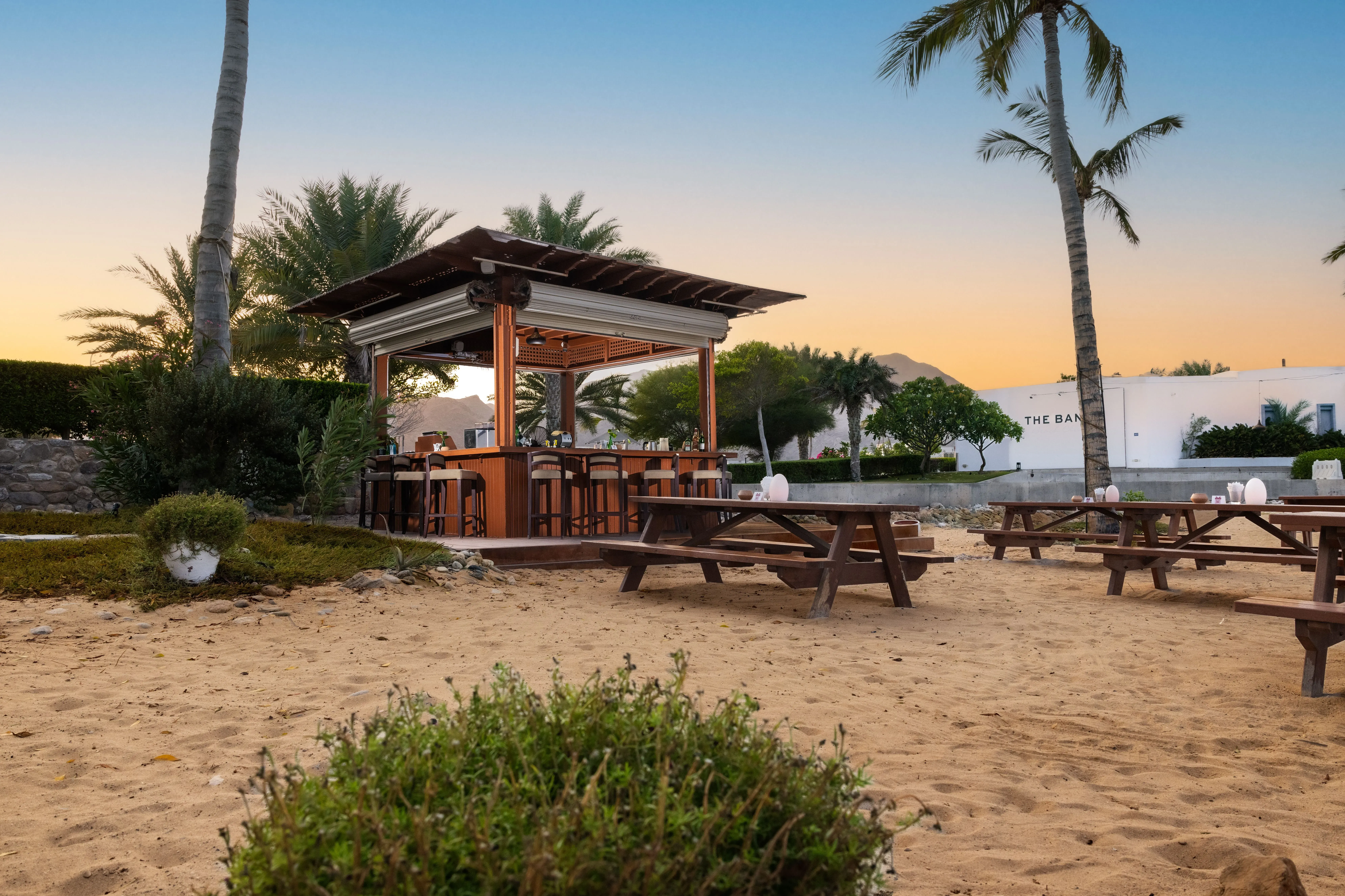 Beach bar with rustic wooden tables, surrounded by palm trees and sandy beach at sunset.