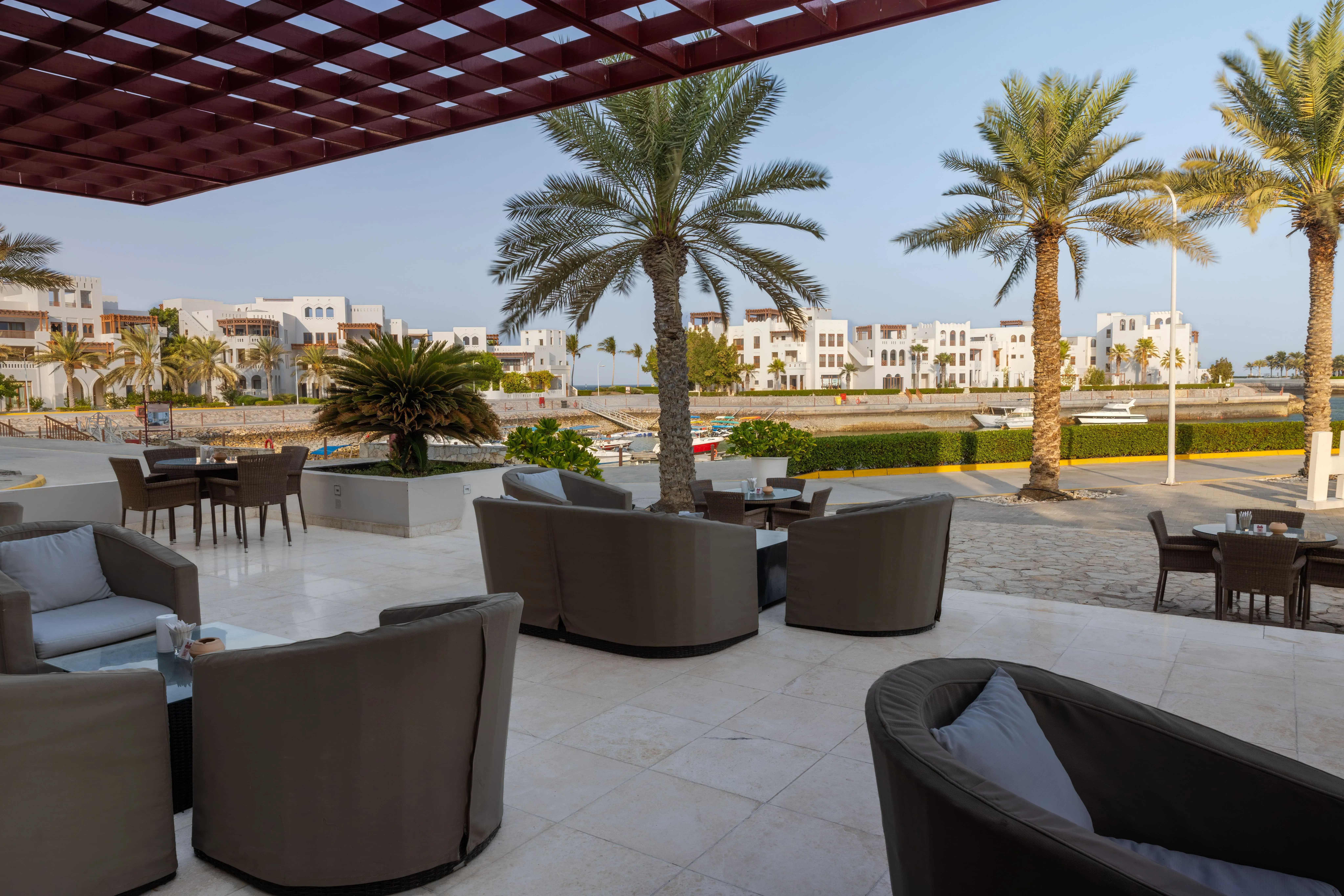 Outdoor lounge area with curved sofas and palm trees, overlooking a waterfront and modern buildings.