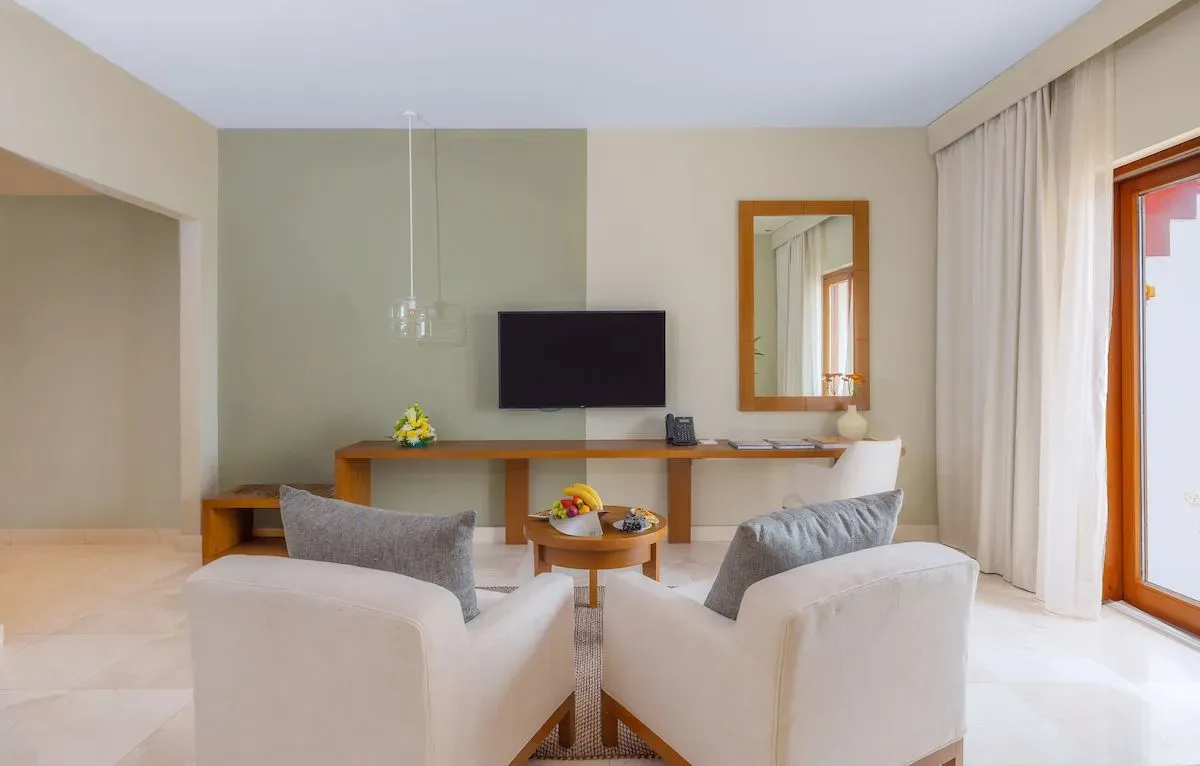 Cozy seating area in a hotel suite featuring two white armchairs, a wooden TV stand, and a window with sheer curtains.