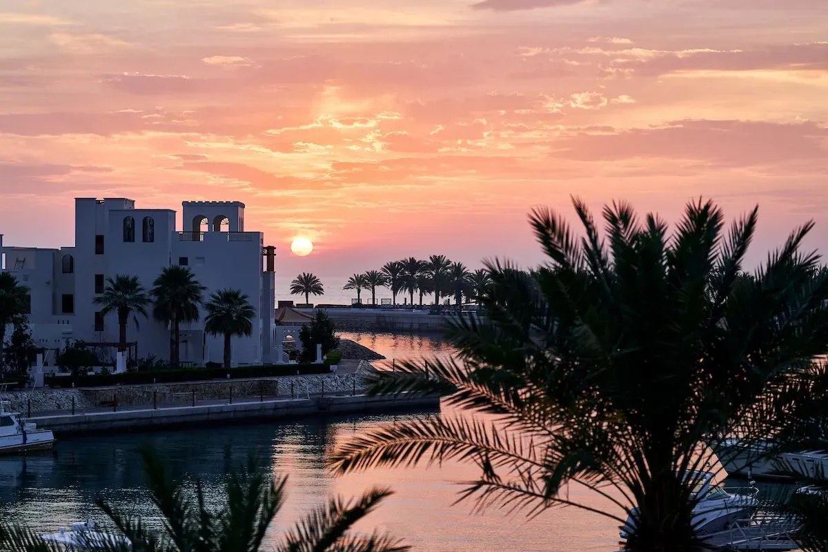 Sunset over the Sifawy Boutique Hotel in Jebel Sifah, with palm trees and a tranquil harbor reflecting vibrant colors.