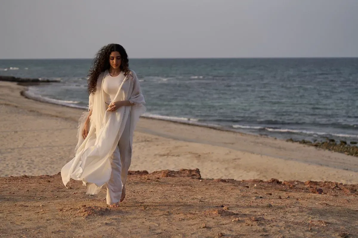 A woman in a flowing white outfit walks along a sandy beach with gentle waves in the background.